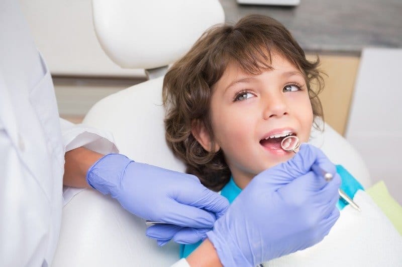 Klinik koltuğundaki çocuk ağzını açmış, hekim ayna ile dişlerine bakıyor