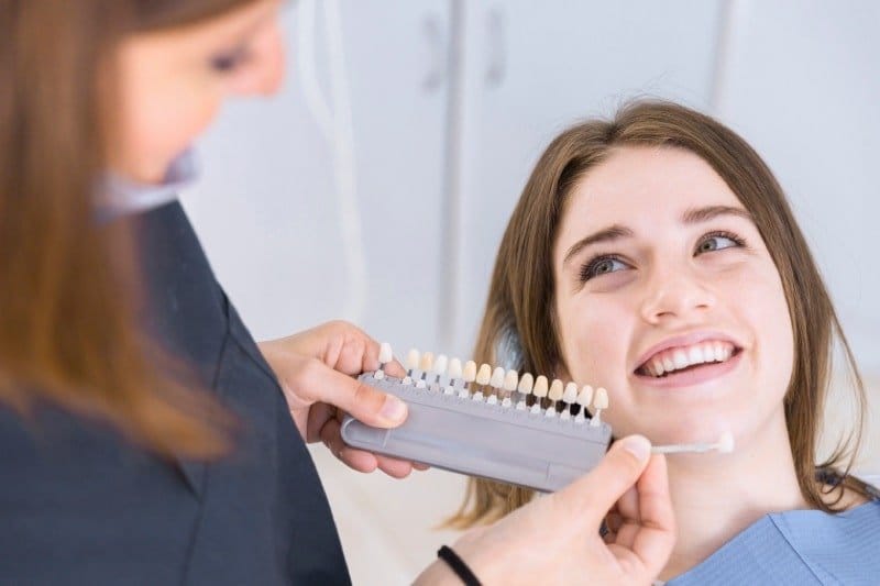 Klinik koltuğunda gülümseyen hastaya diş renk skalası karşılaştırma için tutuluyor